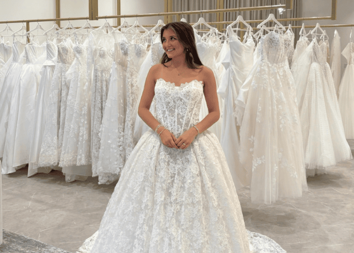 A woman in a white lace wedding gown stands smiling in a bridal shop, surrounded by dresses. The atmosphere is joyful and elegant.