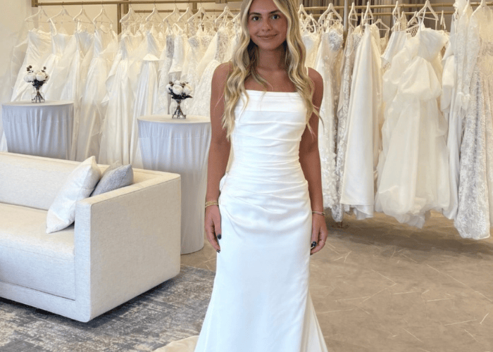 A woman in a sleek white wedding dress stands in a bridal boutique, surrounded by hanging gowns. The ambiance is elegant and serene.