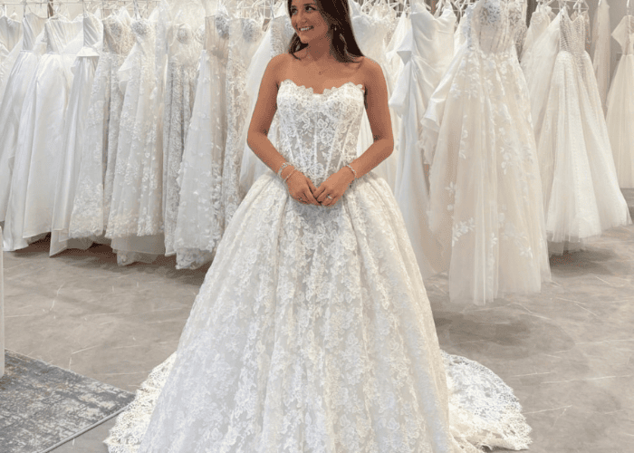 A woman in a bridal shop tries on a lace wedding gown with a strapless, corset-style bodice. She stands smiling, surrounded by other dresses.
