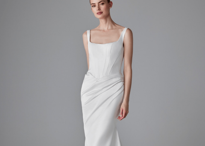 A woman in a sleek, elegant white gown stands against a gray background. The dress features a square neckline and draped details, exuding sophistication.