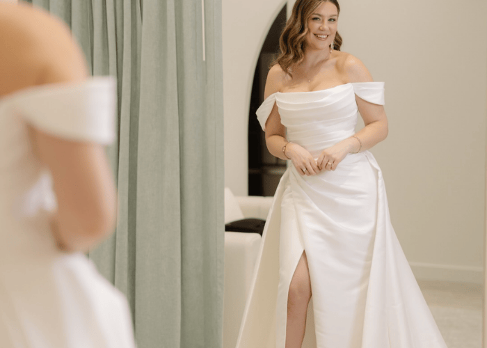 Woman smiling in a bridal boutique, wearing an off-shoulder white wedding dress with a front slit. Soft lighting and an elegant, joyous atmosphere.