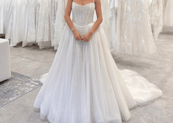 A woman in an elegant, beaded wedding gown stands in a bridal boutique surrounded by other dresses. The gown’s intricate details convey a sense of sophistication and joy.