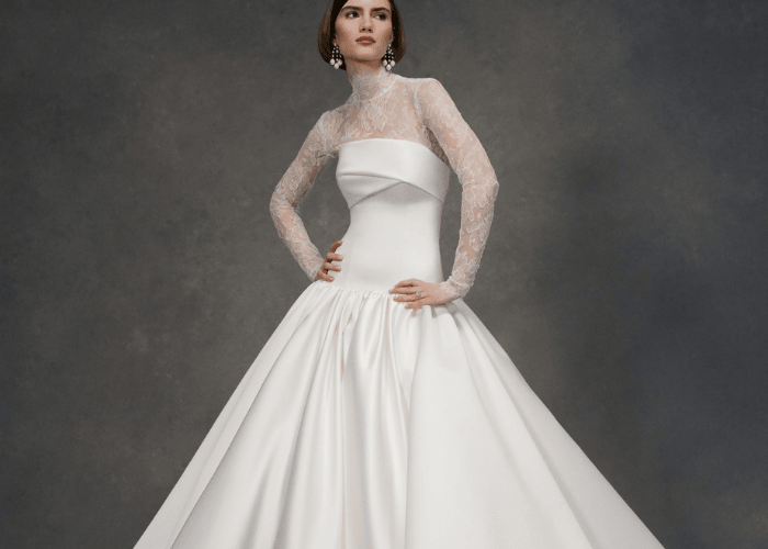 A model poses confidently in an elegant white gown with a full satin skirt and lace long-sleeve bodice. The backdrop is a muted gray, enhancing the gown's sophistication.