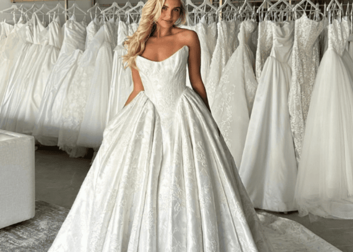 A bride in a strapless, basque waist full-skirted wedding gown poses in a bridal shop, surrounded by elegant white dresses. The mood is serene and graceful.