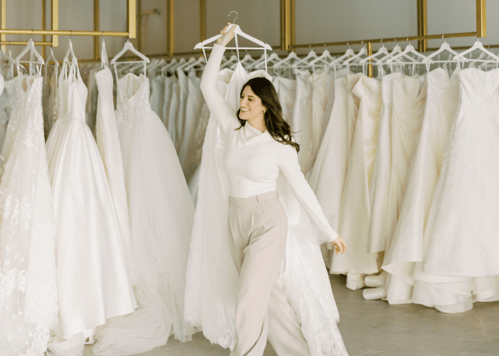 What to Wear Wedding Dress Shopping (And What to Avoid) (1) A smiling woman twirls with a wedding dress on a hanger in a bridal shop. She stands among rows of elegant white gowns, exuding joy and excitement.