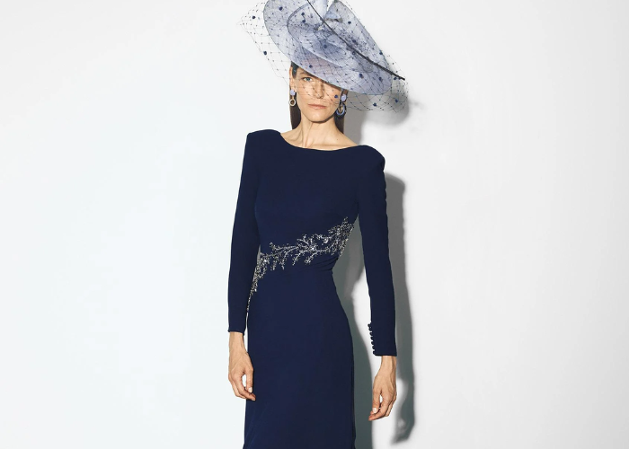 Navy blue mother of the bride dress with long sleeves, embellished waist detail, bateau neckline, and statement fascinator.