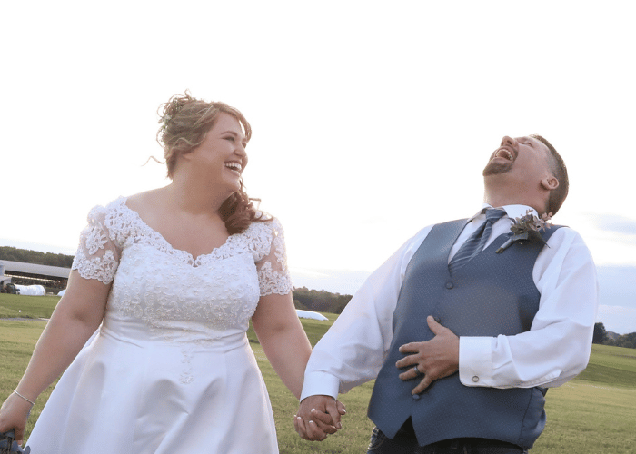 Plus size bride and groom laughing together outdoors after their wedding ceremony, with the bride wearing a lace wedding dress with short sleeves.