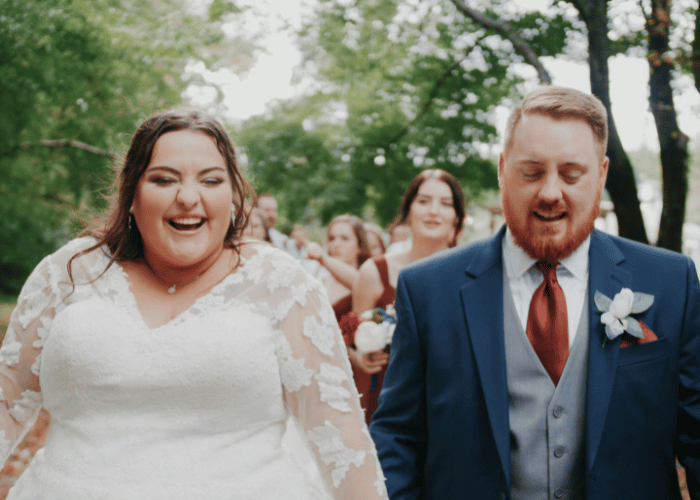 Smiling plus size bride wearing a long sleeve lace wedding dress walking with the groom after their wedding ceremony.