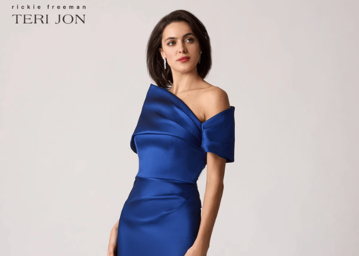 Woman wearing a royal blue satin off-the-shoulder dress with sculptural draped detailing across the bodice